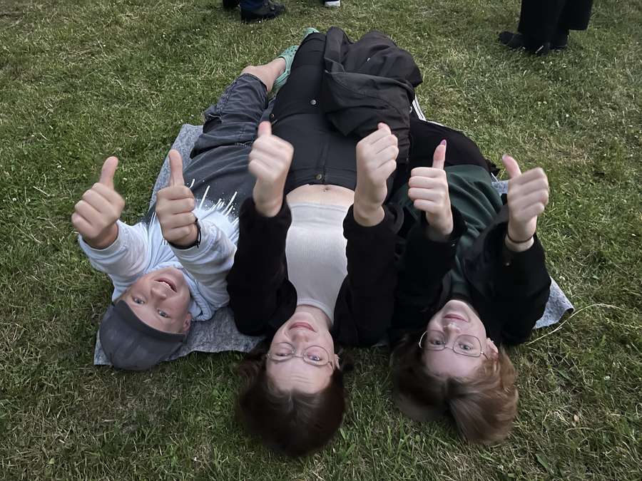 Maise, Laura, and Elliot lying in the grass, giving thumbs up